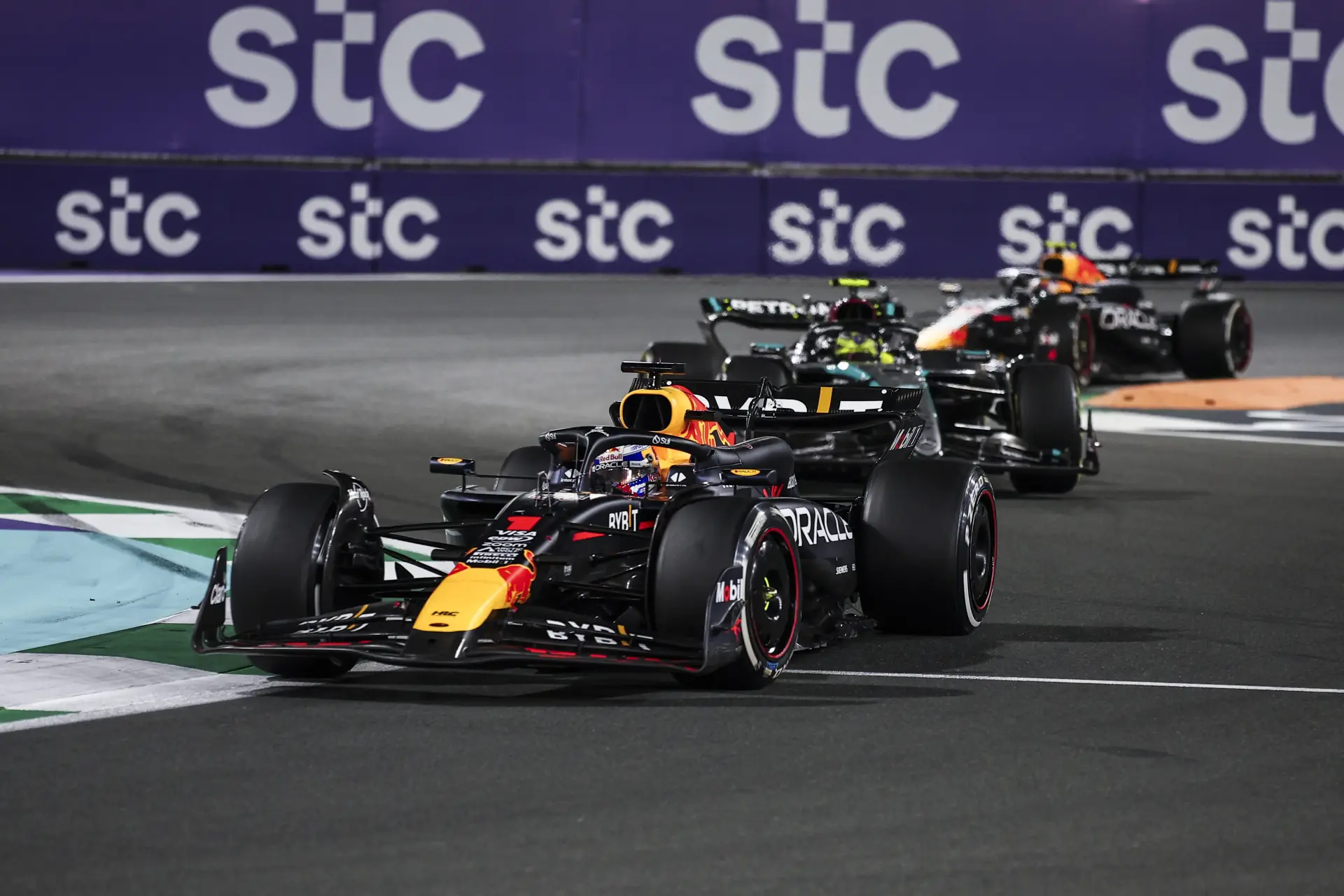 Red Bull Racing Formula 1 car leading a Mercedes and another Red Bull car through a corner during the Saudi Arabian Grand Prix, with "stc" sponsorship banners in the background.