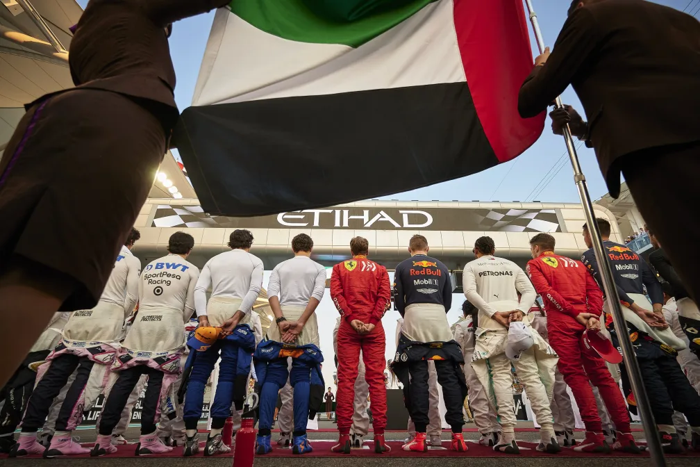 Formula 1 drivers standing in a row before the Abu Dhabi Grand Prix, viewed from behind under a large UAE flag, with Etihad branding in the background.