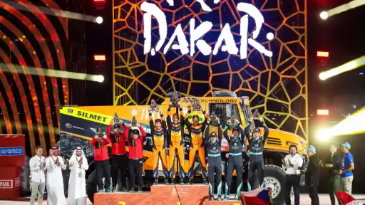 Dakar Rally truck podium ceremony with winning teams celebrating and holding trophies in front of a large Dakar Rally logo and an orange race truck.