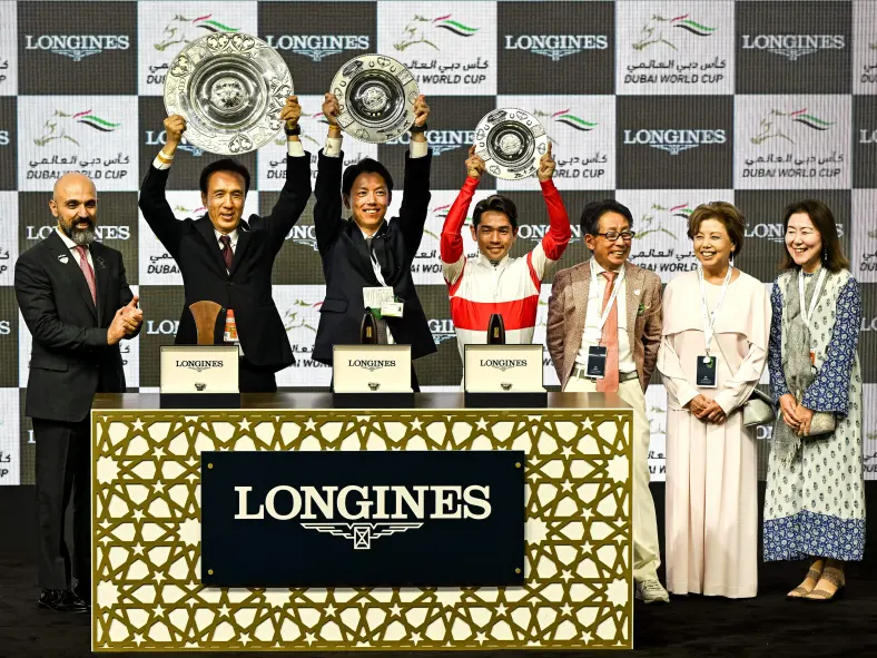 A professional award ceremony scene with individuals holding up trophies, a backdrop displaying the Longines and Dubai World Cup logos, and a decorated podium area, focusing on event sponsorship.