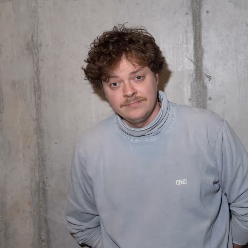 Junger Mann mit lockigem Haar und Schnurrbart trägt hellgrauen KITH-Pullover vor Betonwand.