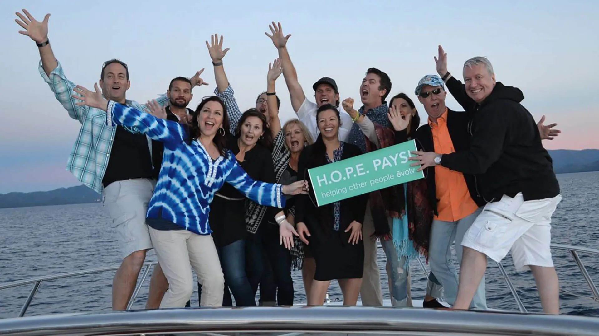 Excited team on boat holding H.O.P.E. PAYS sign with mountains backdrop