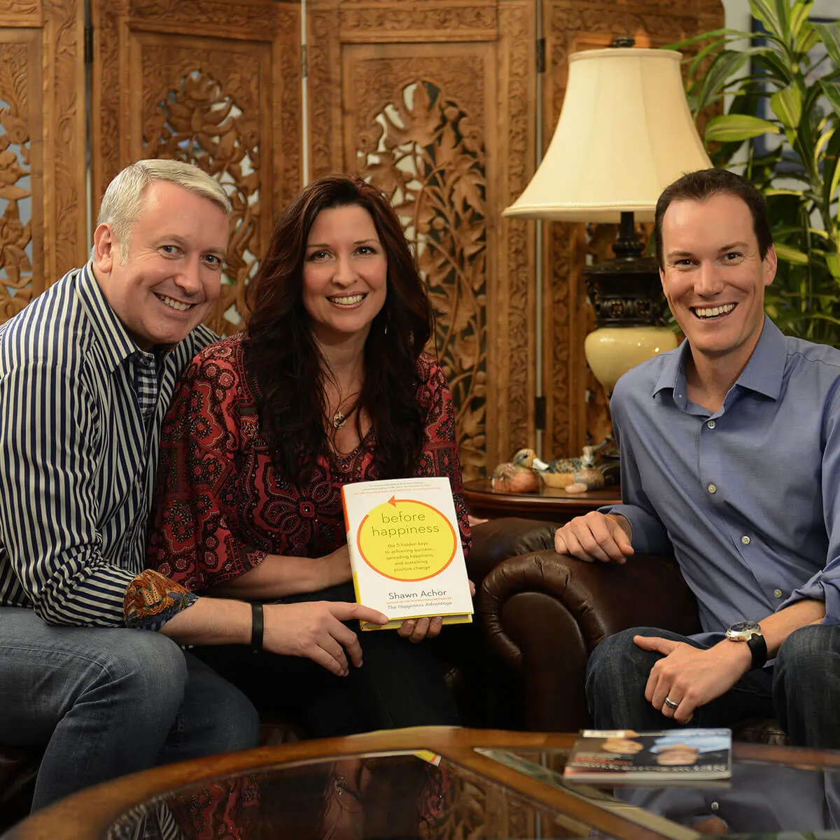 Three people smiling, holding "Before Happiness" book in cozy setting