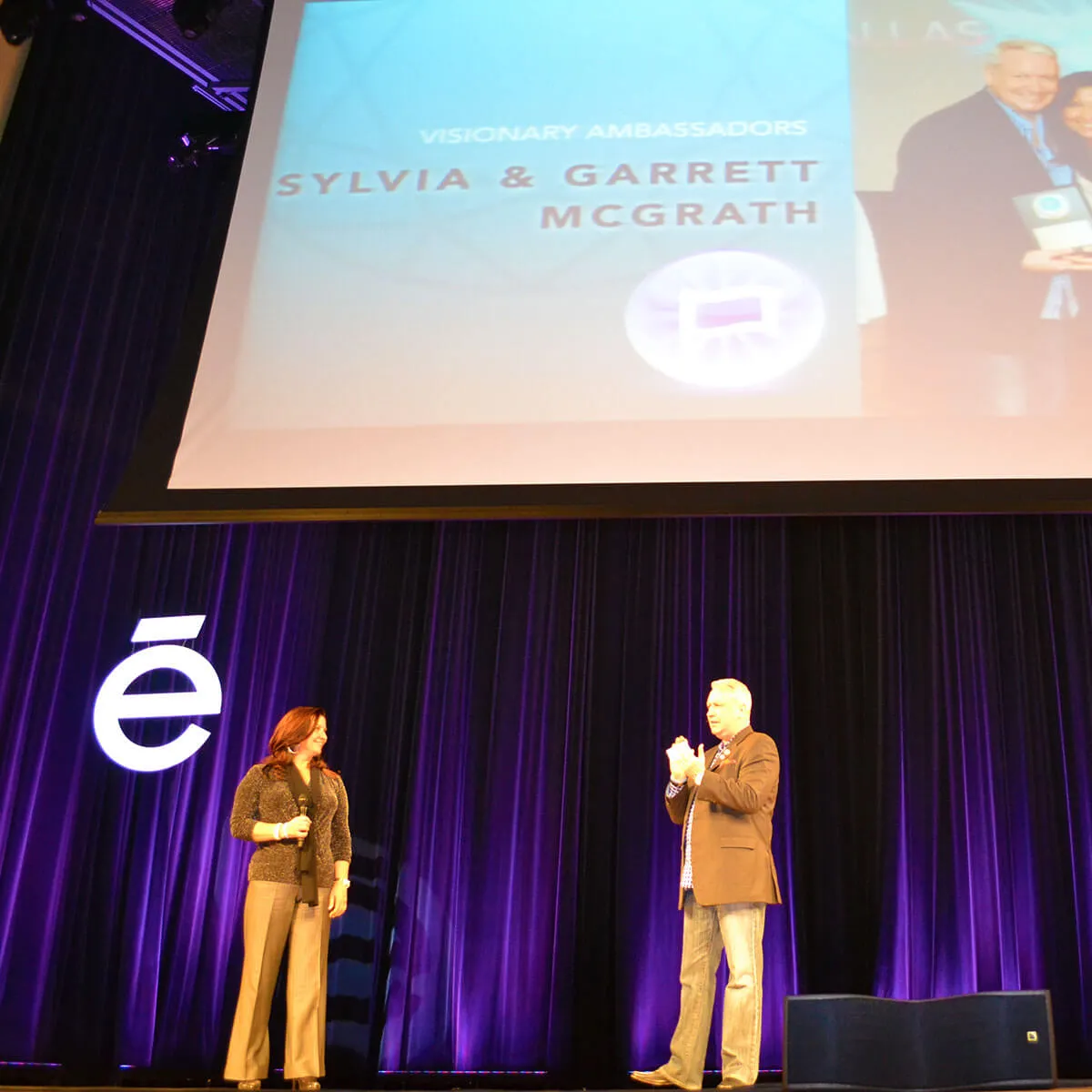 Two speakers on stage at conference with Visionary Ambassadors slide