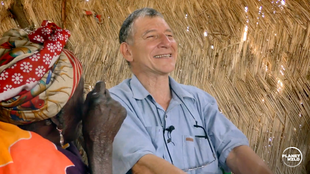 A man in a light blue shirt sits smiling inside a structure with thatched walls. A small dark-colored dog or puppy sits on his lap, and colorful decorative items are visible on the wall behind him.