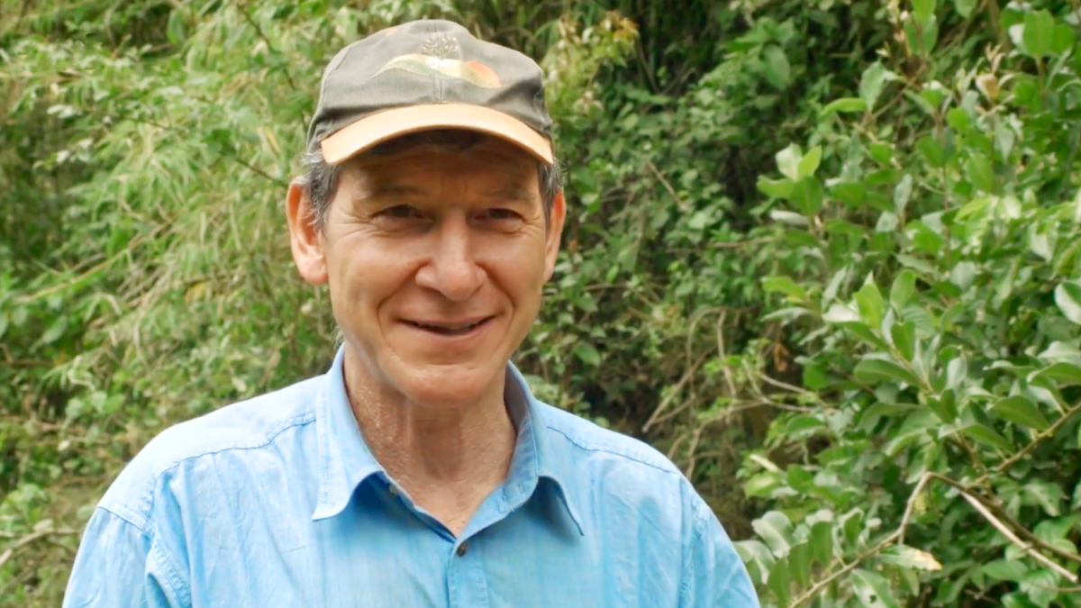 Tony Rinaudo wearing a light blue shirt and an olive-green cap smiles at the camera while standing in front of dense green vegetation and tropical foliage.