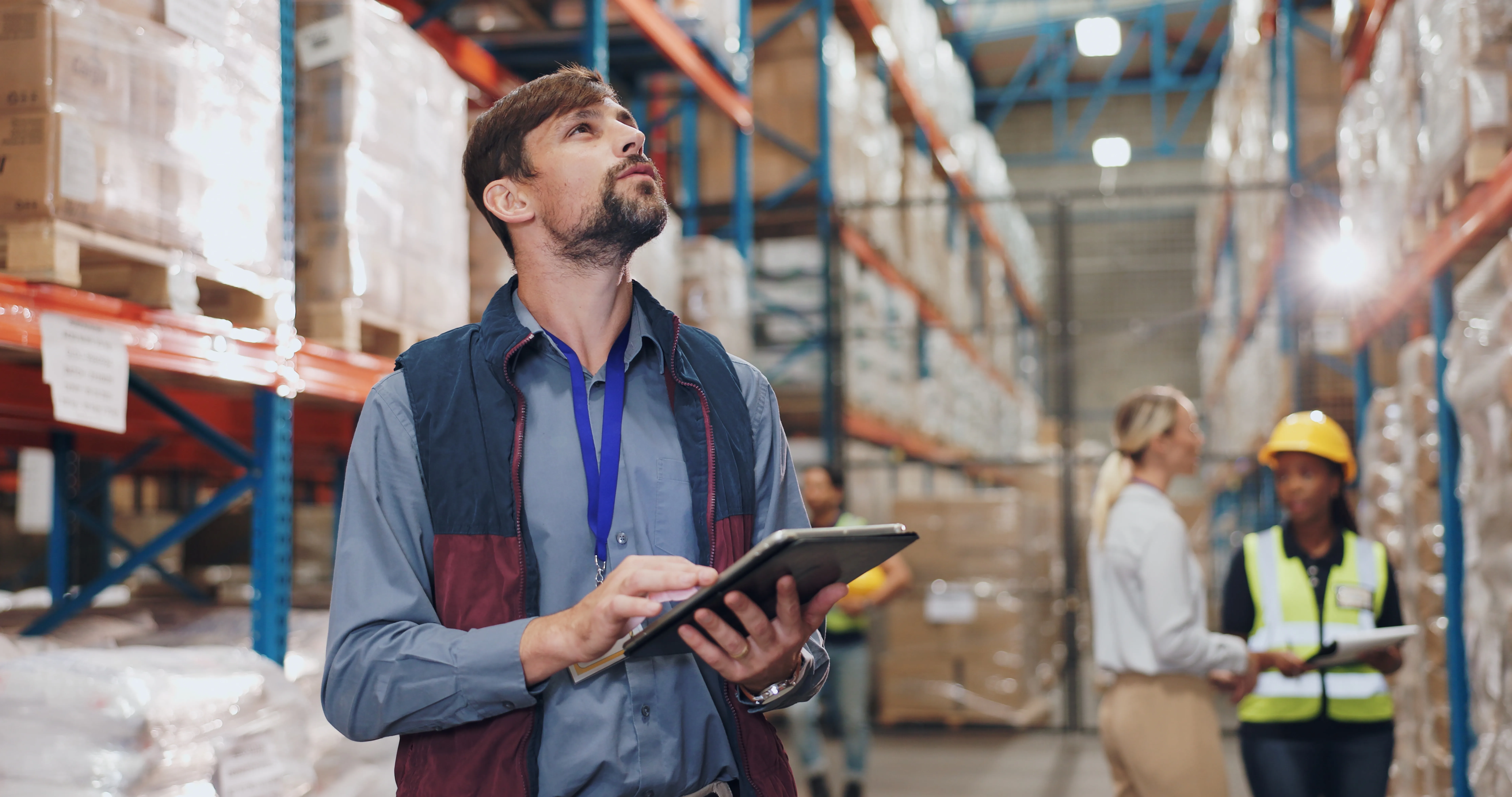 Man with tablet looking up while standing in a warehouse with workers and stacked boxes around.