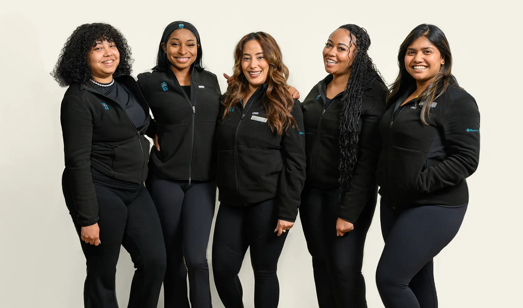 A diverse group of five women standing closely together in black zip-up jackets and black pants, smiling at the camera against a plain light background.