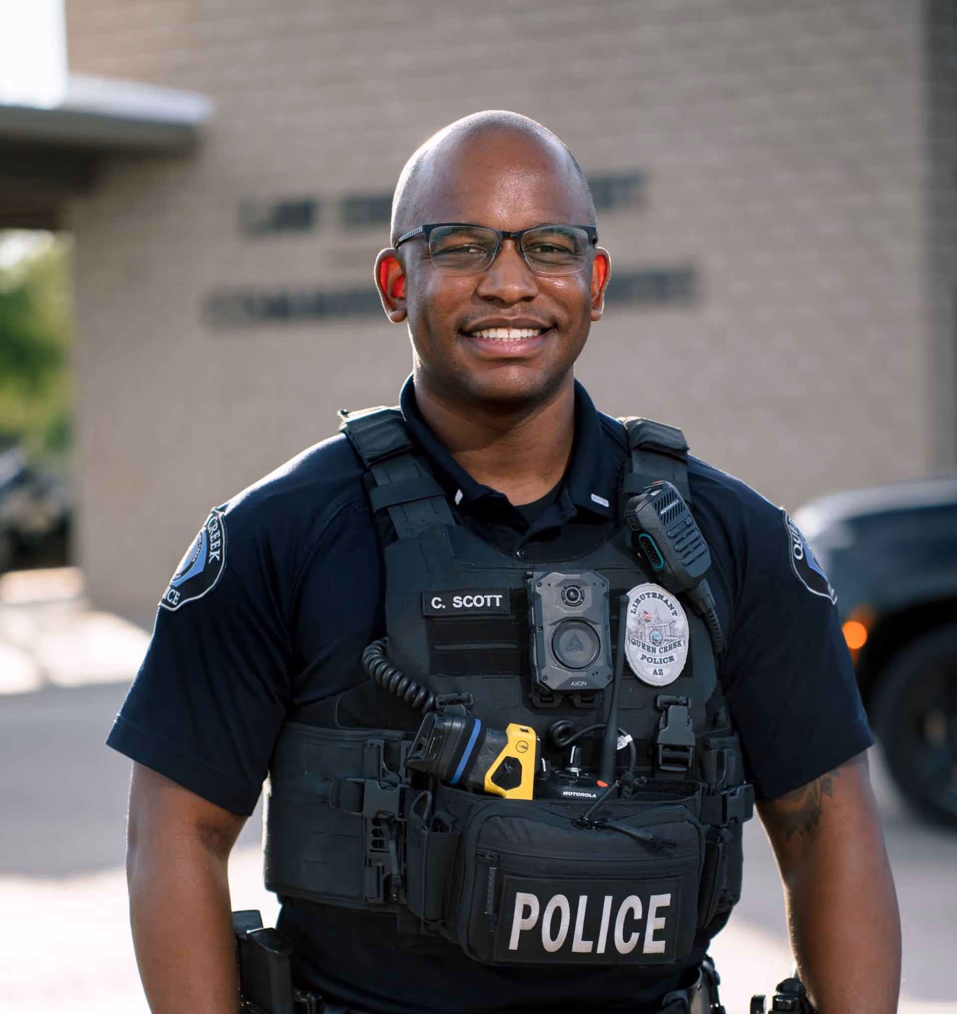 Smiling male police officer wearing glasses and tactical vest labeled.