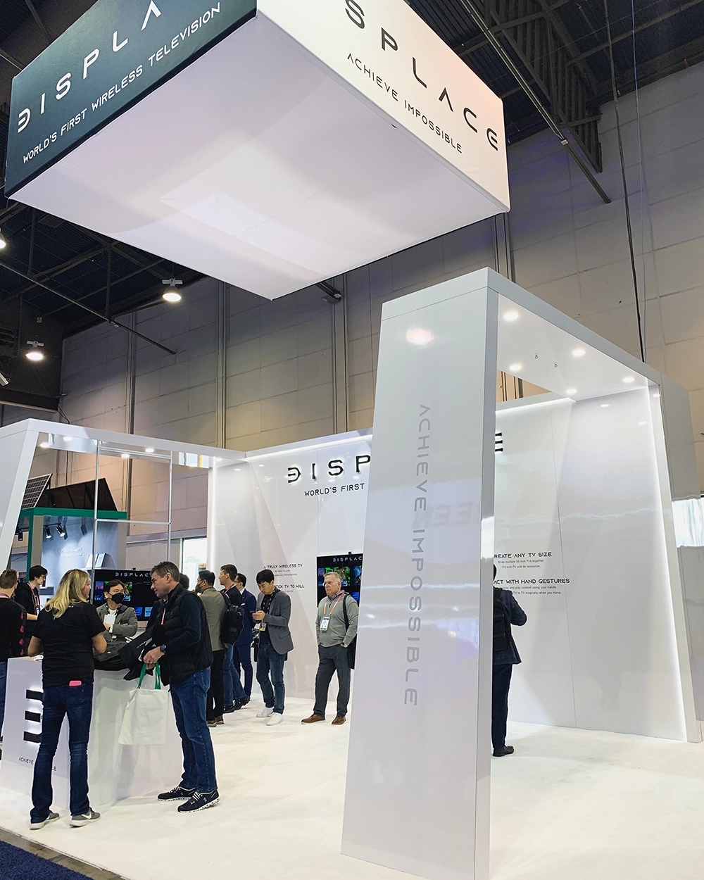 Trade show booth with a white modern design displaying 'Displace' and 'Achieve Impossible' with several people interacting inside.