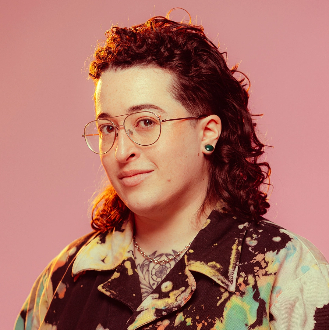 A picture of EJ, a white nonbinary person with a curly brown mullet and wire-rimmed glasses.