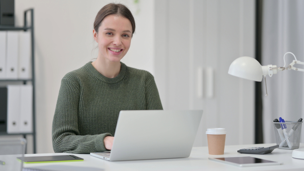 Vrouw met groene trui die glimlacht en op een laptop werkt aan een witte bureau met koffiebeker en bureaulamp.