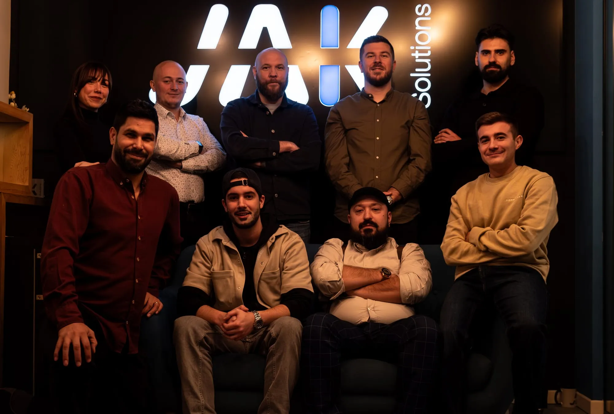 Group photo of nine casually dressed colleagues posing in front of a black wall with a partially visible illuminated company logo.