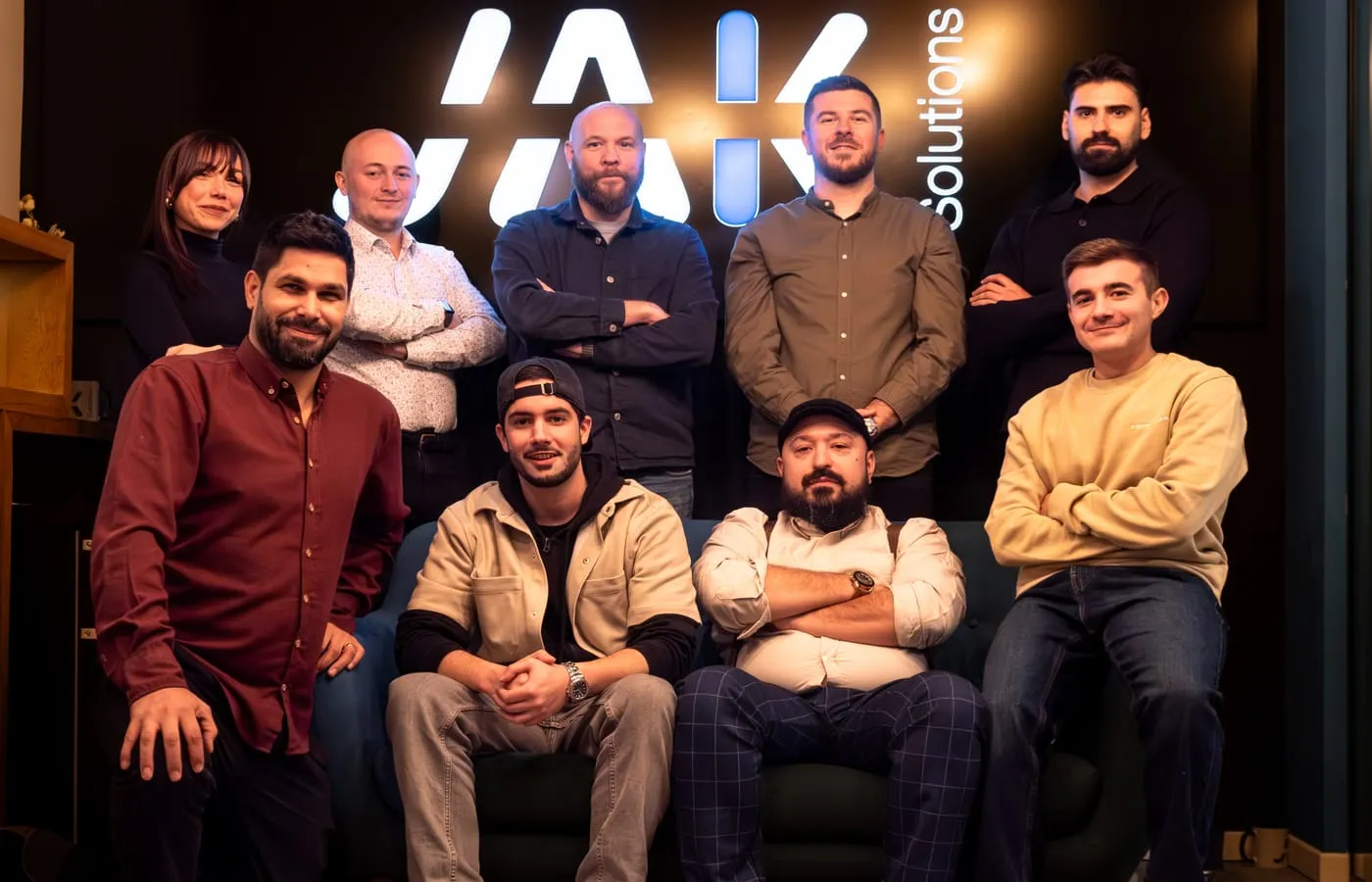 Group of nine casually dressed people posing indoors in front of a lit company logo, with some seated on a couch and others standing.
