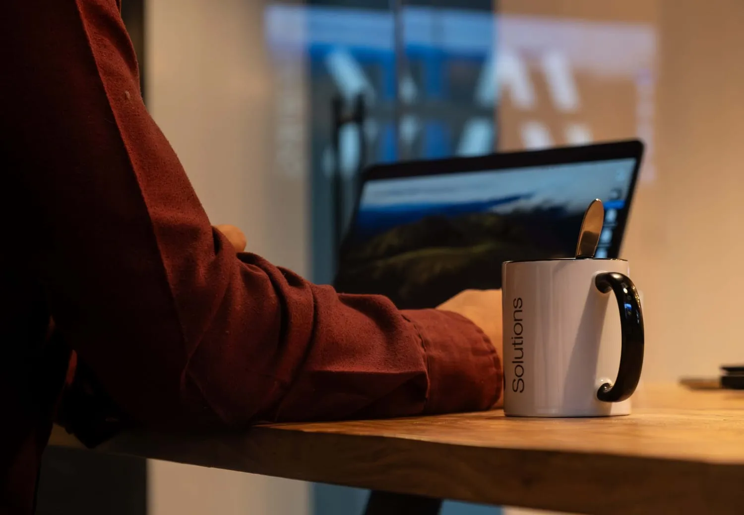 Person in a maroon shirt sitting at a wooden table with a white mug labeled 'Solutions' and an open laptop in the background.