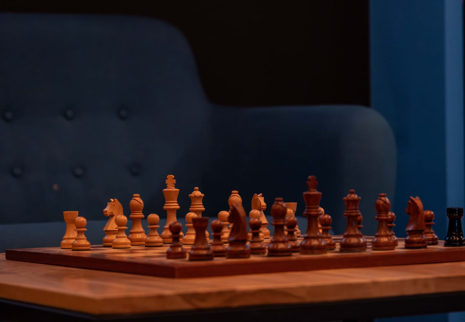 Wooden chessboard set up for a new game with pieces in starting positions on a table in front of a dark blue cushioned chair.