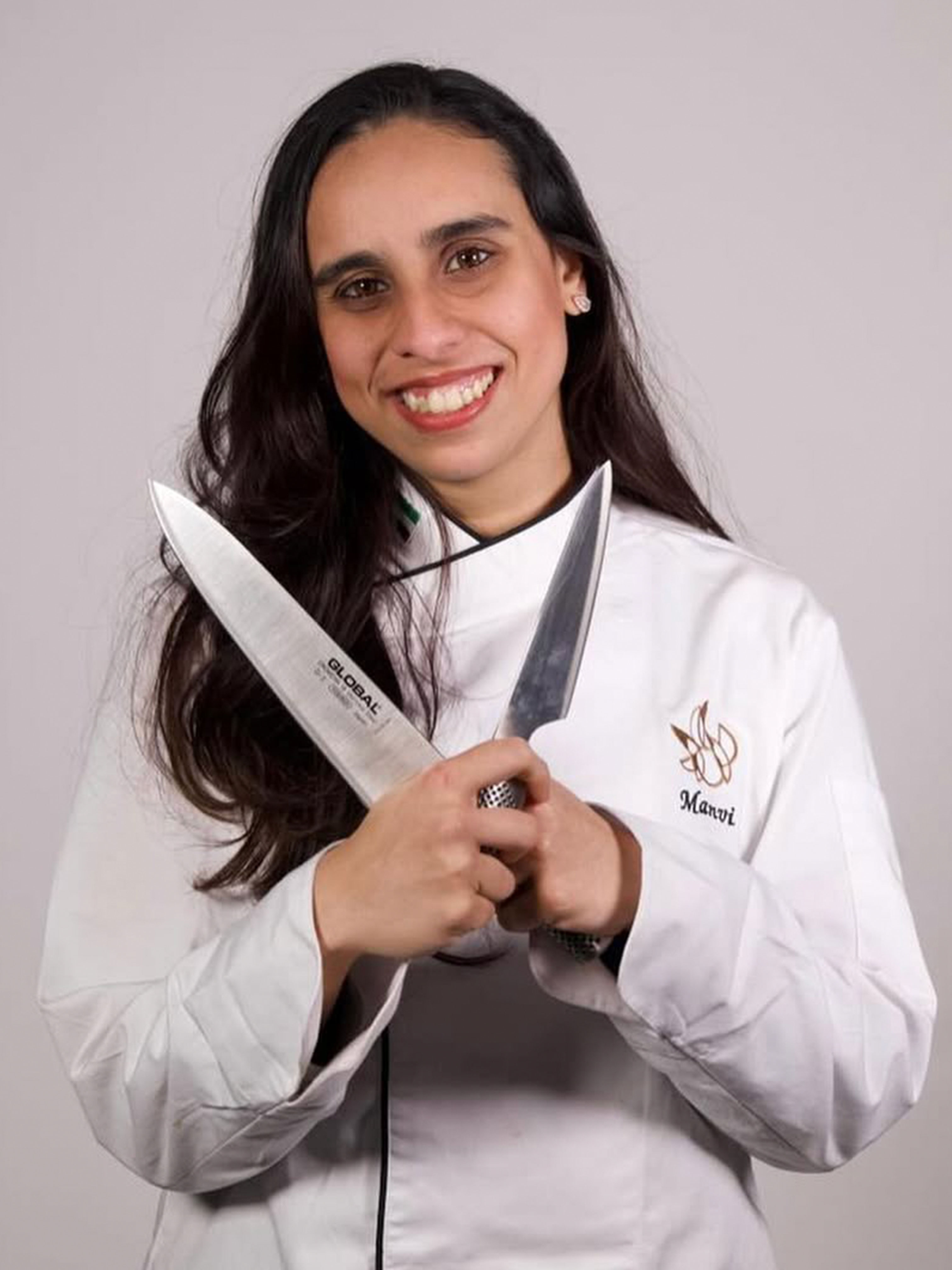 Smiling woman in white chef jacket holding two kitchen knives crossed in front of her.