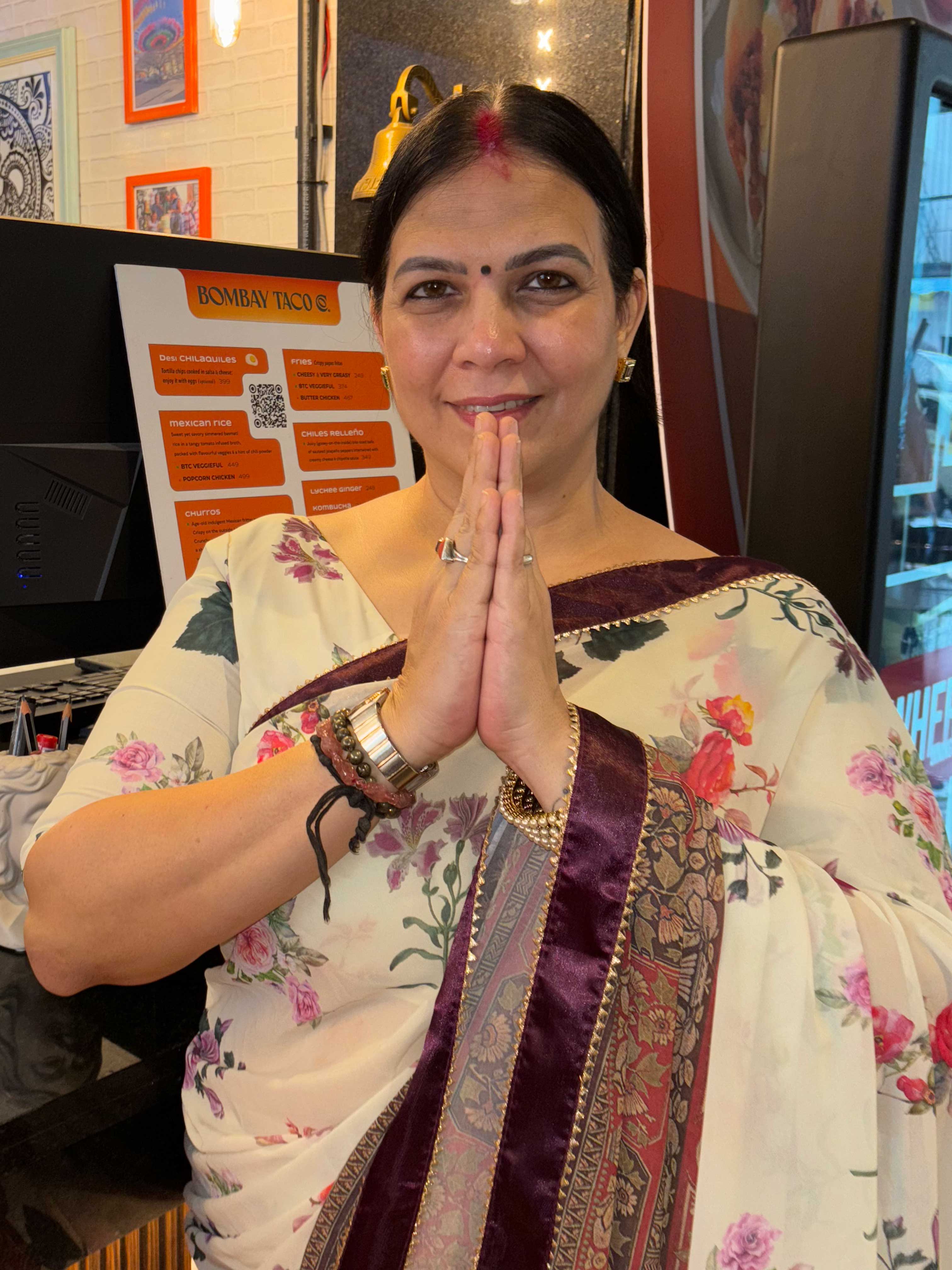 Woman wearing a floral saree with a maroon and gold border, smiling and holding hands together in a traditional greeting gesture.