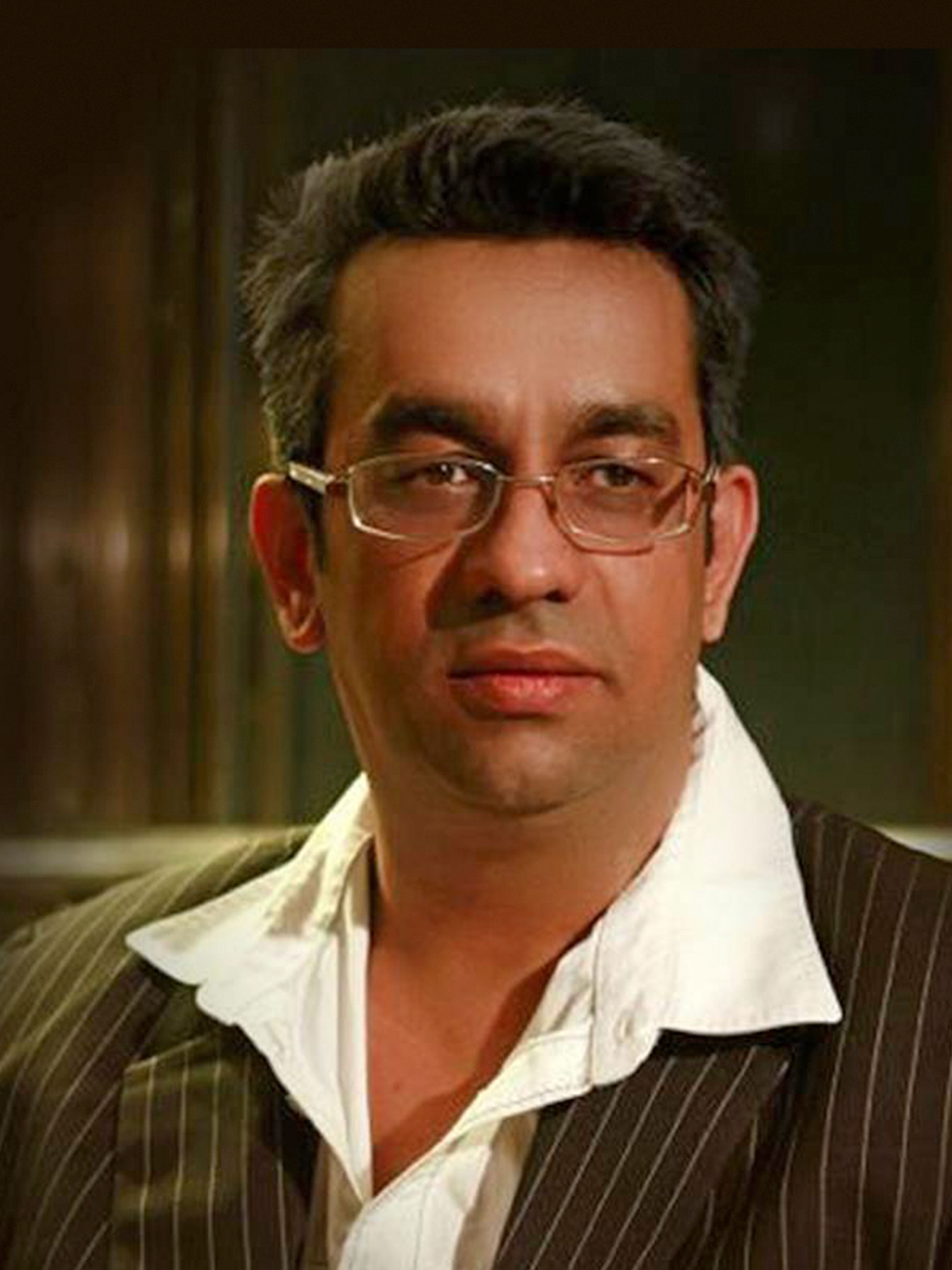 Middle-aged man wearing glasses and a white shirt under a pinstripe jacket in a dimly lit room.