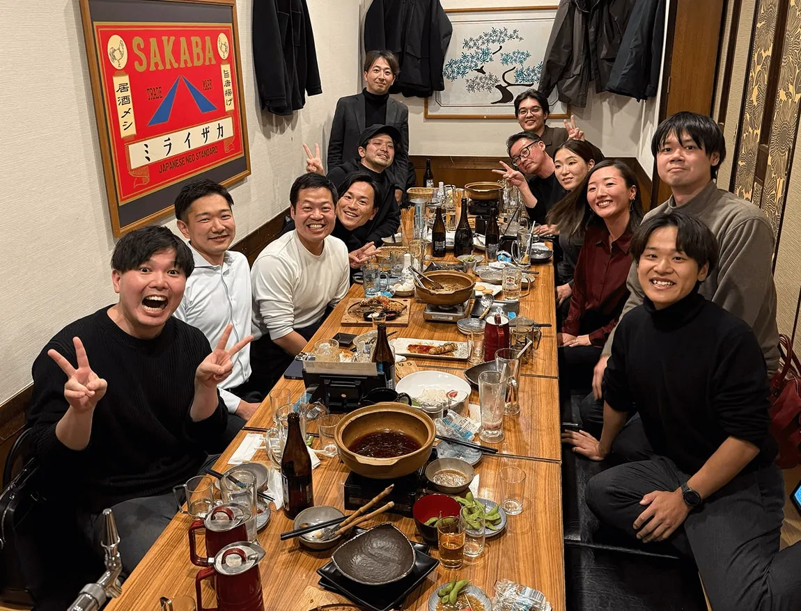 A group of twelve people smiling and posing with peace signs around a long wooden table filled with food and drinks in a cozy restaurant.