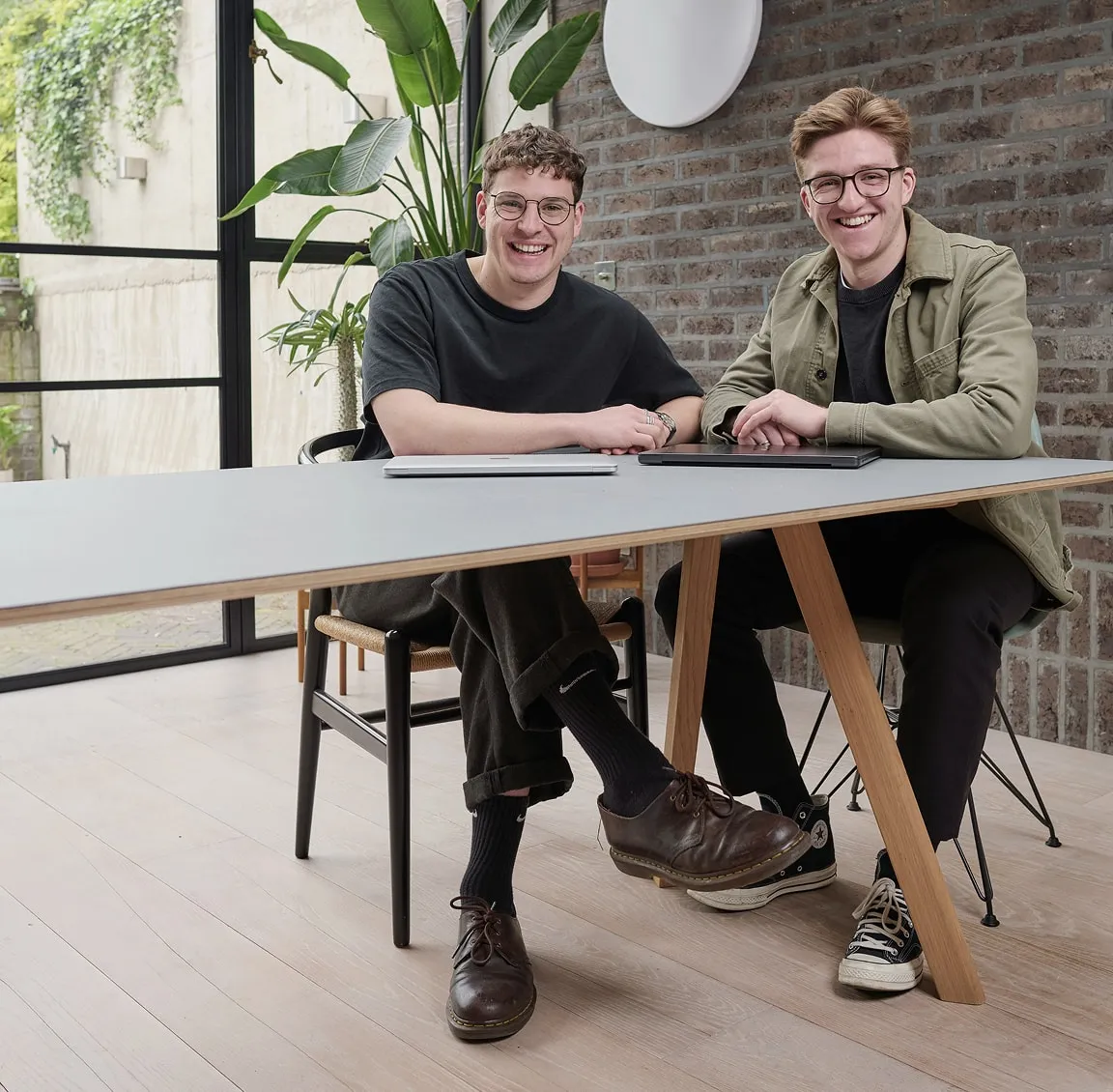 Two young men sitting at a modern table, smiling, each with a laptop in front of them in a bright room with plants and a brick wall.