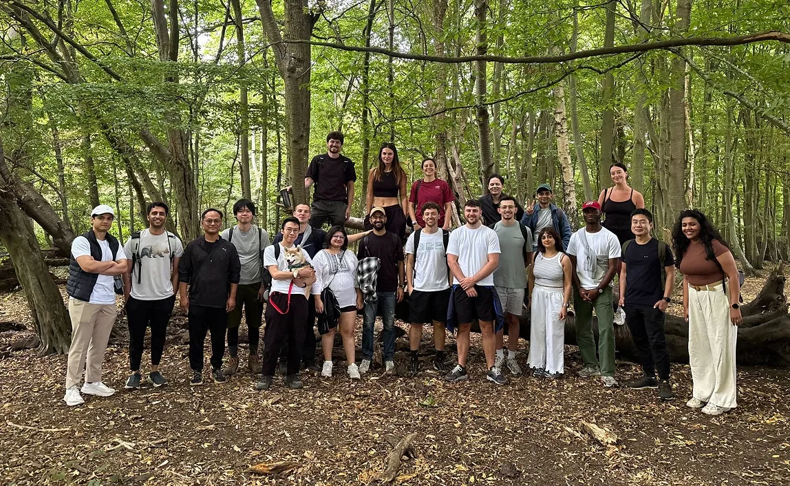 Group of twenty people and a dog posing in a green forest with trees and fallen leaves on the ground.