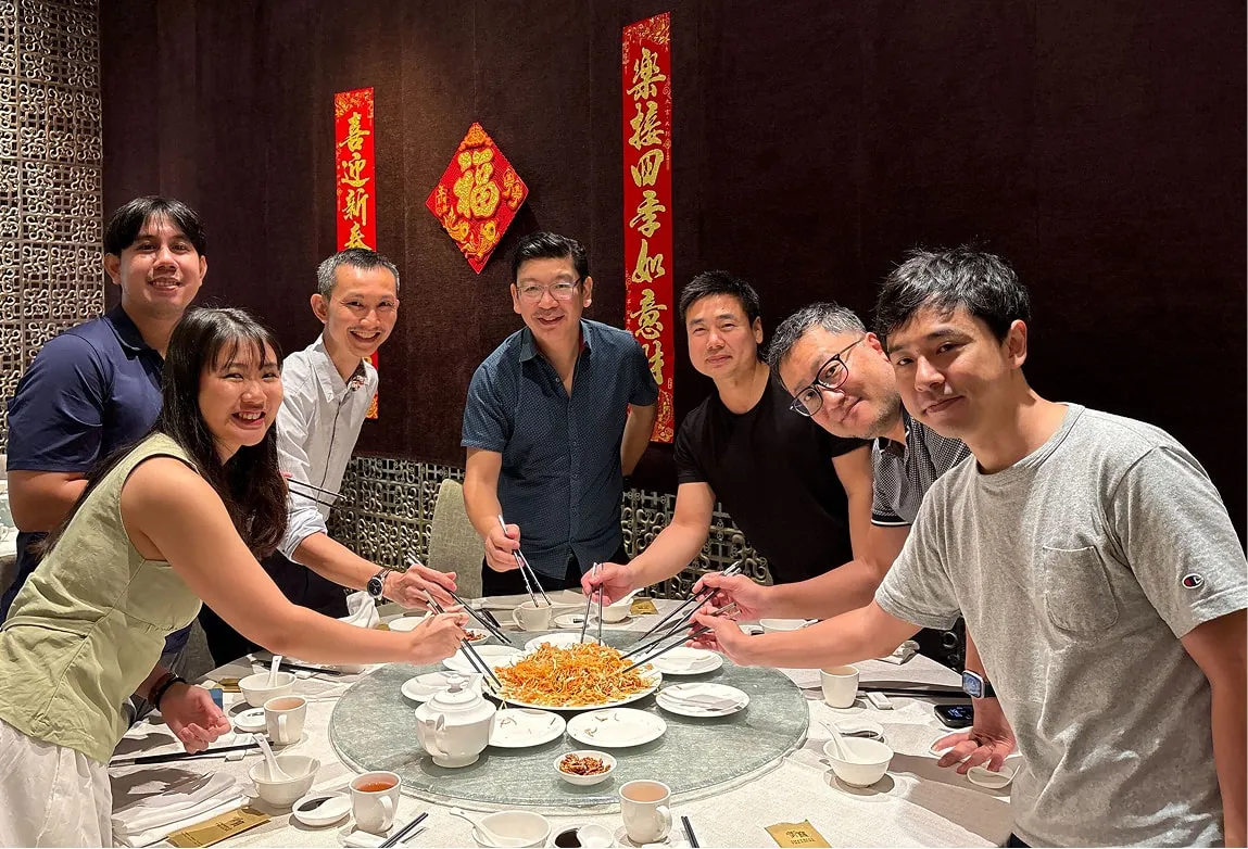 Seven people around a round dining table sharing a plate of noodles with chopsticks in a restaurant with Chinese New Year decorations on the wall.