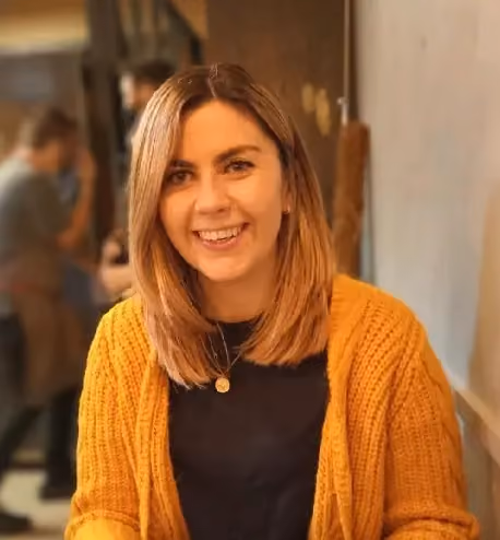 Smiling woman with shoulder-length hair wearing a mustard sweater and black top in an indoor setting.
