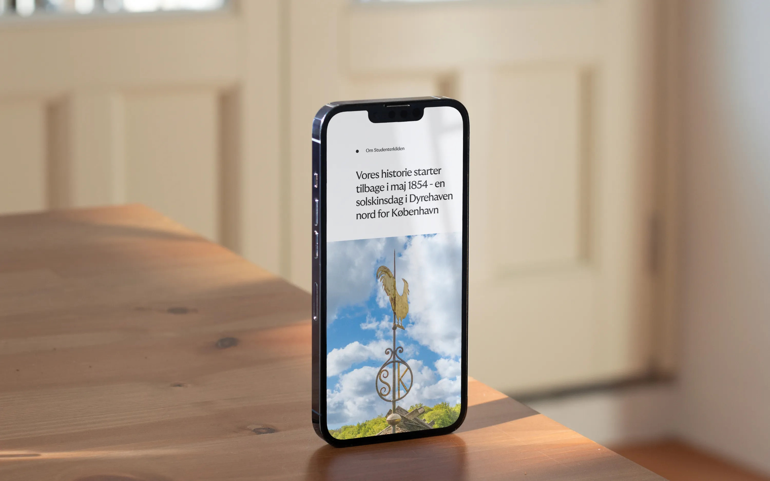 Smartphone on table showing Danish text and a weather vane with a rooster against a cloudy sky.