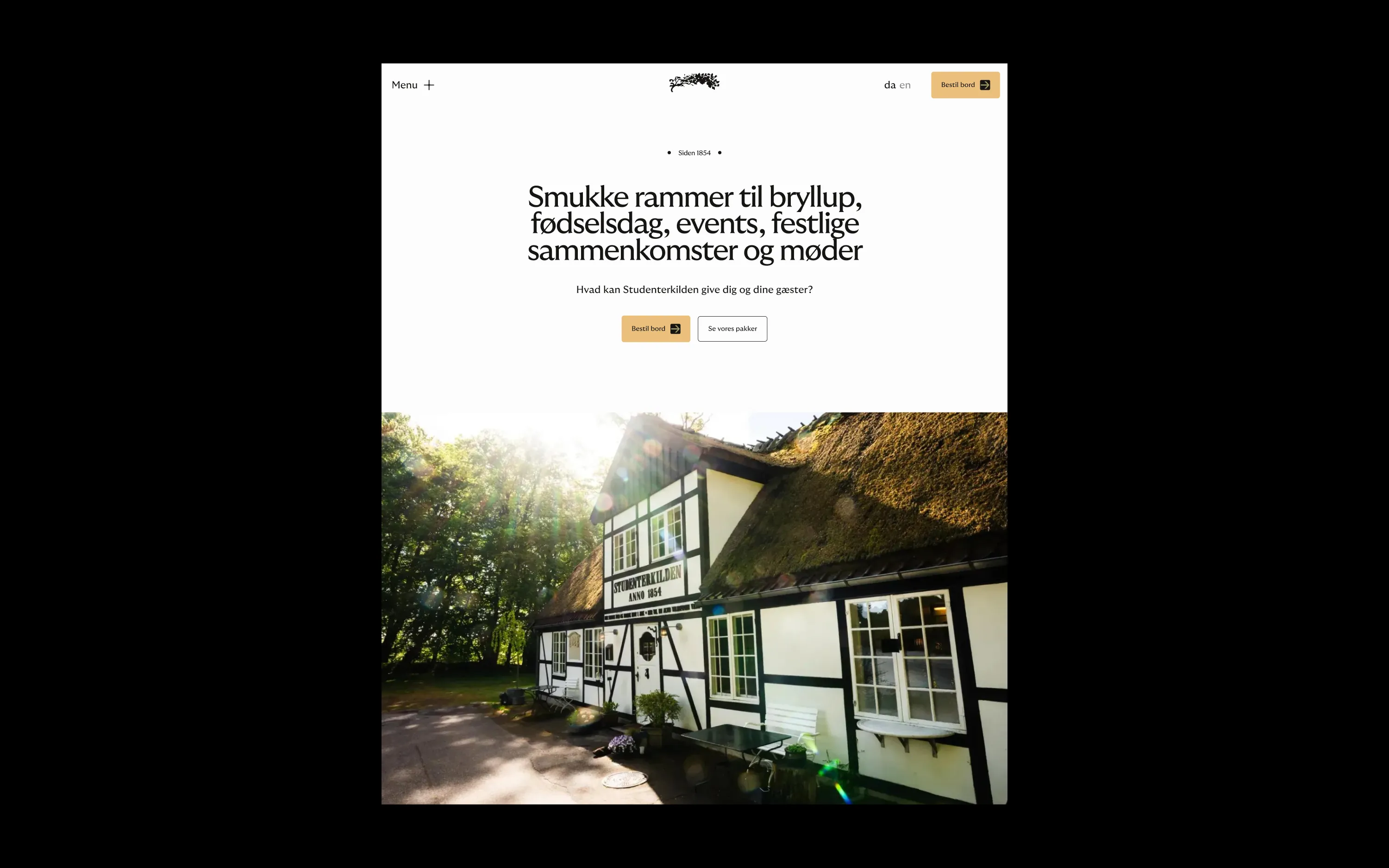 White and black timber-framed house with a thatched roof in sunlight surrounded by trees.