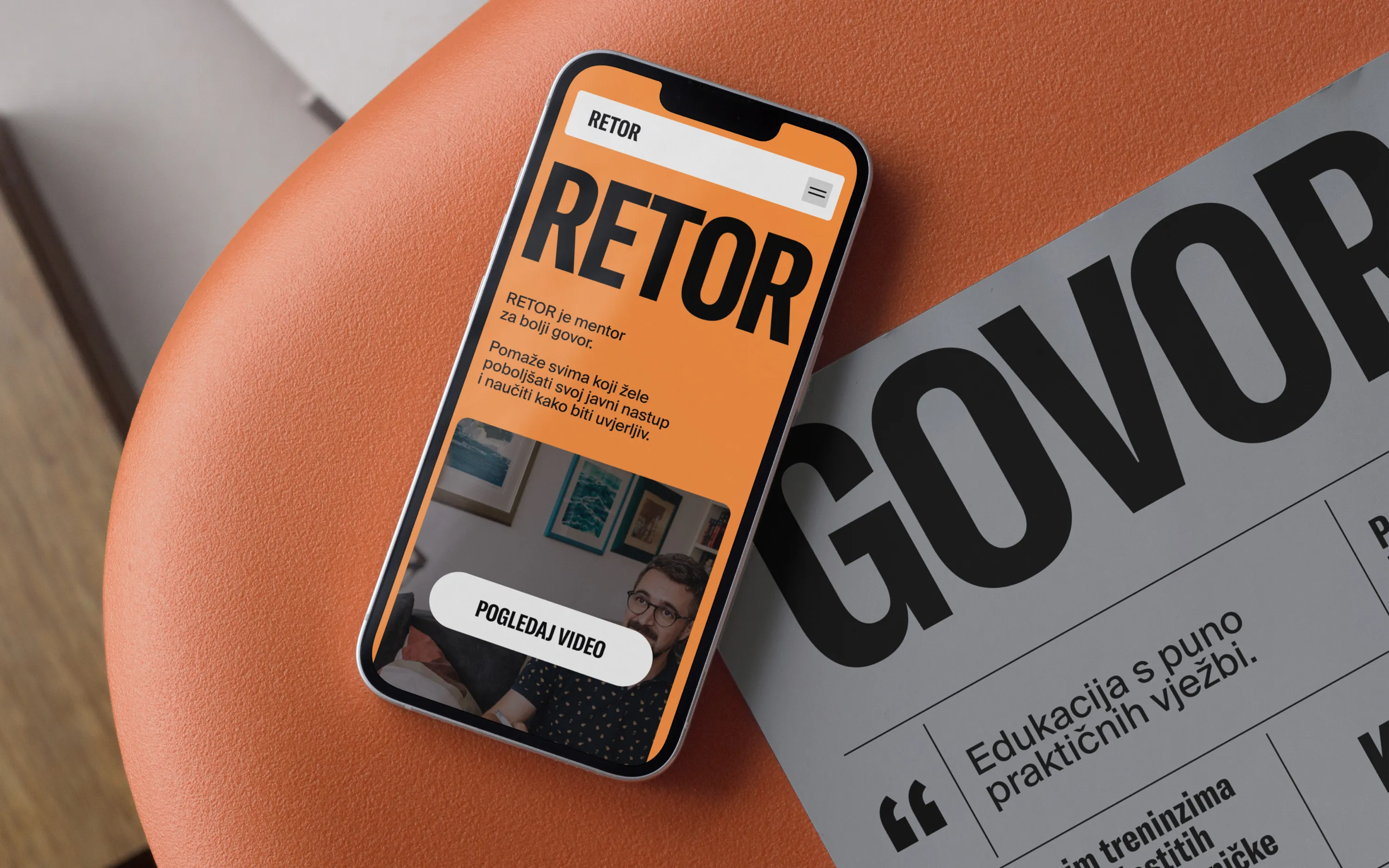 Smartphone on orange chair showing RETOR mentor website with video button and text in Croatian.