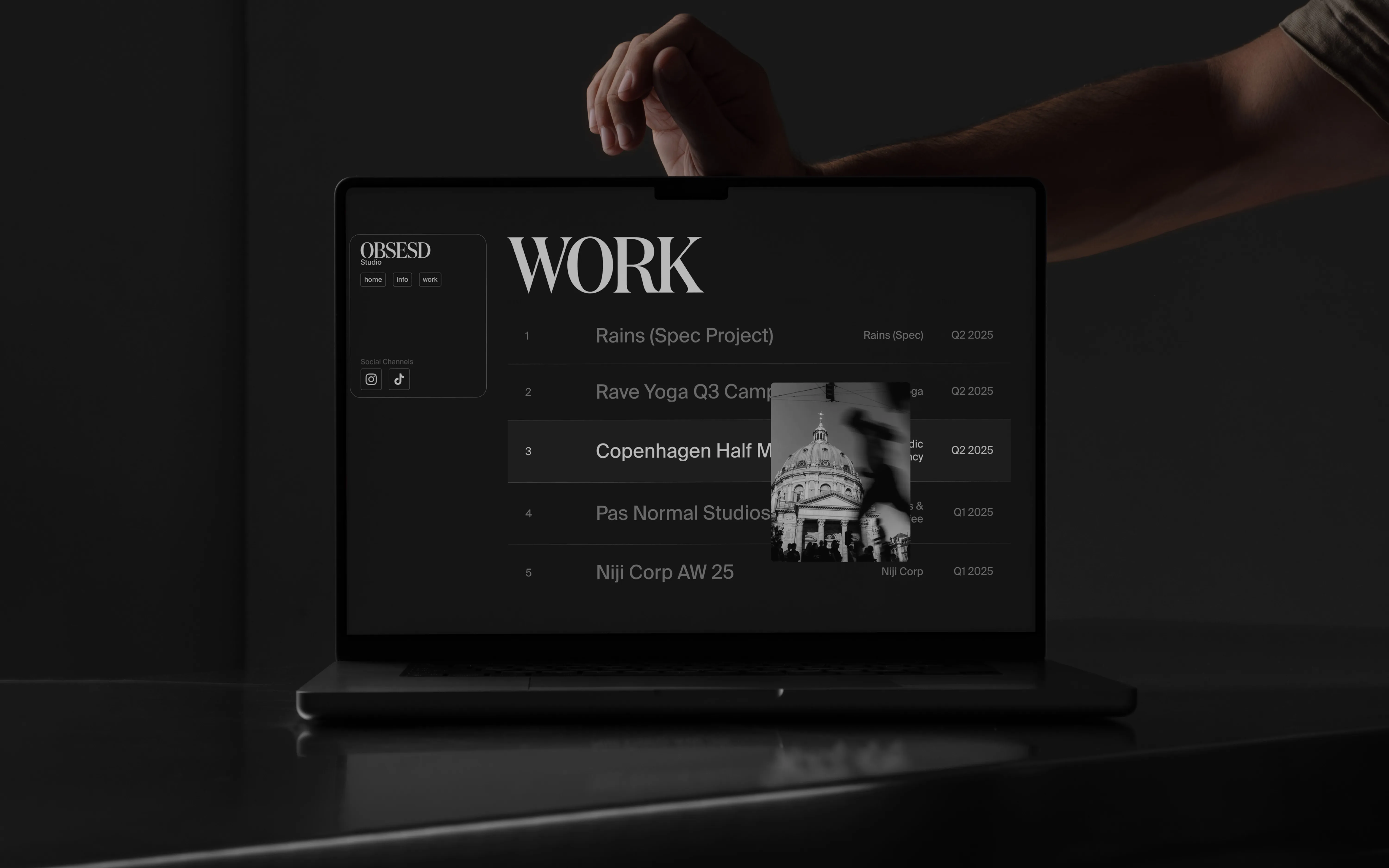 Laptop screen showing a work list on a website with project titles and a black-and-white building photo overlay.
