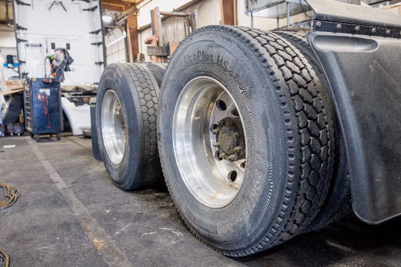Close-up of dual semi-truck tires inside repair shop, showing detailed tread patterns, metal rims, and workshop equipment in background.