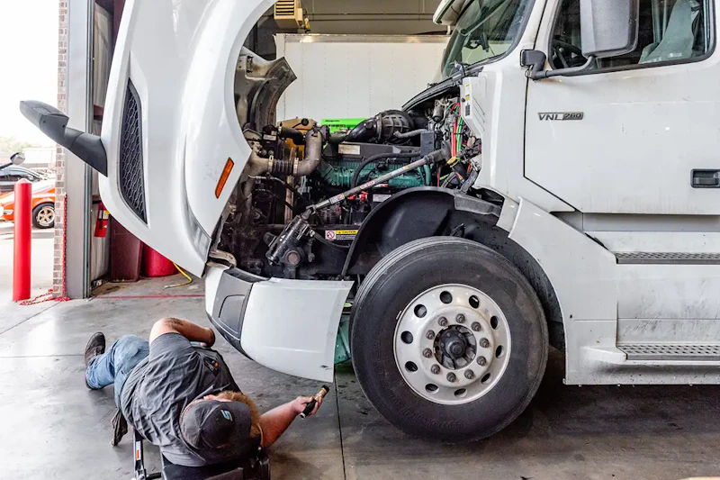 DOT inspection with technician under Volvo VNL 760 checking front-end components and engine bay for safety compliance.