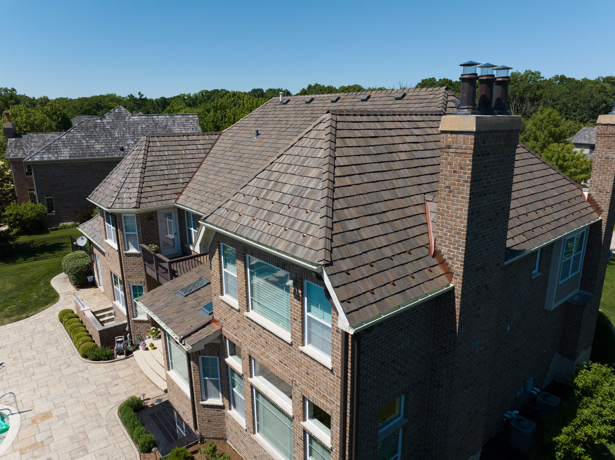 Large brick house with multiple gable roofs, chimneys, and a spacious paved driveway with greenery.