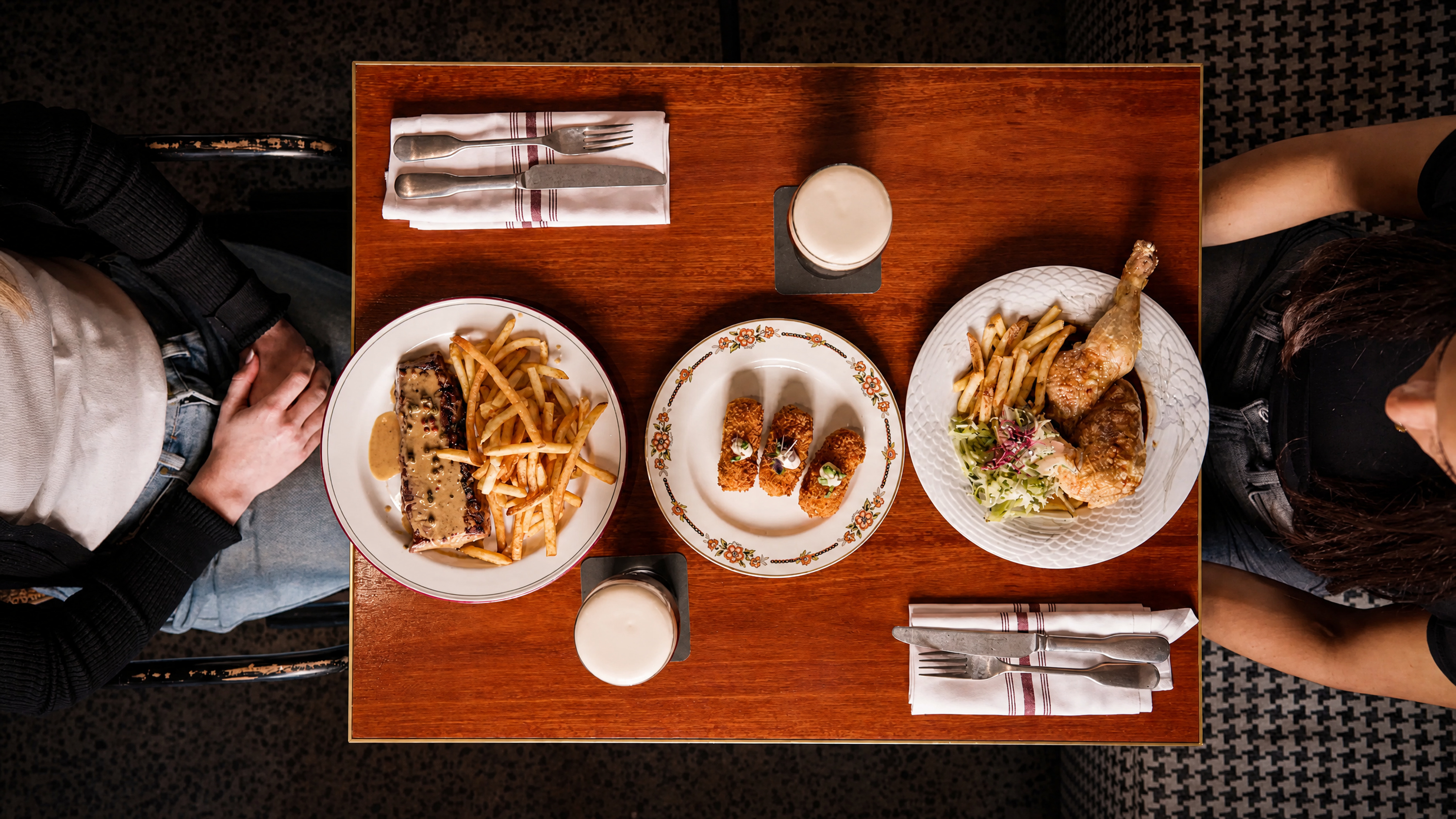 Top view of a wooden table set for two with plates of food: left plate has steak with sauce and fries, center plate has three fried croquettes with toppings, right plate has roasted chicken drumsticks, fries, and salad; two drinks with foam are also on the table, each place setting includes folded napkins with knife and fork.