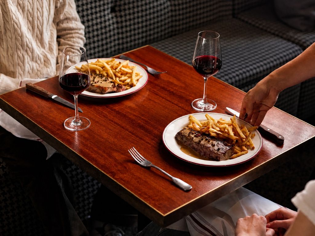 Two plates on a wooden table each with steak, French fries, and a glass of red wine, with one person reaching for fries and another sitting opposite wearing a beige cable knit sweater.