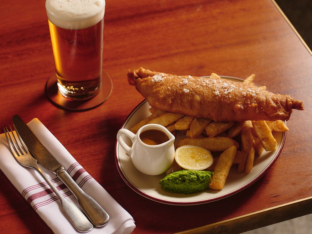 Plate of battered fish and chips with thick fries, a mound of mushy peas, half a lemon, and a small jug of brown sauce on a wooden table next to a glass of beer with foam and cutlery wrapped in a white napkin.