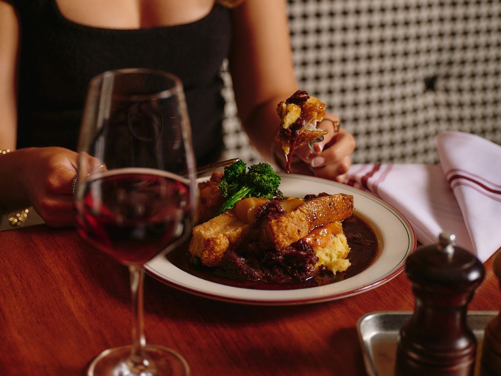 Person wearing a black sleeveless top using a fork and knife to eat the Sunday Roast, a plate of roasted meat, broccoli, and potato on a white plate, with a glass of red wine in the foreground on a wooden table.