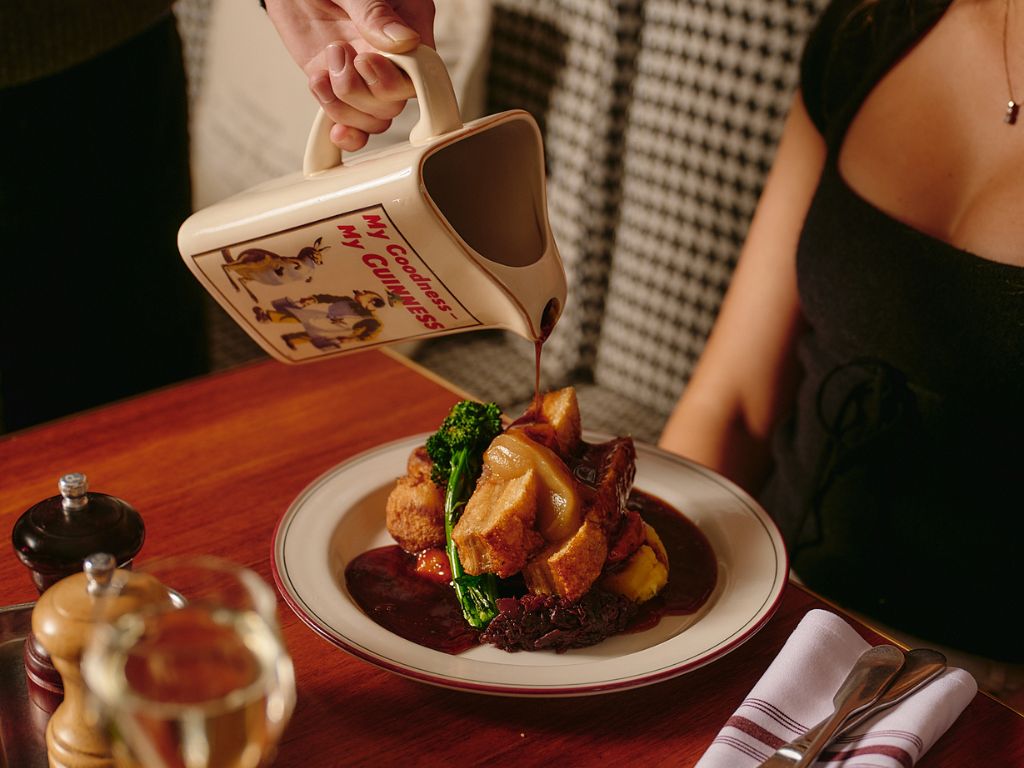 Hand pouring dark gravy from a mug labeled 'My Goodness My Guinness' onto a Sunday Roast plated meal of roasted meat, broccoli, mashed potatoes, and other vegetables at a wooden table with utensils and a glass of water.