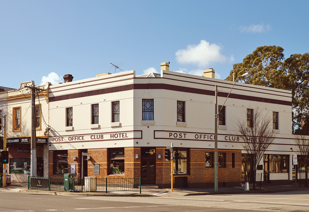 Corner view of a two-story building with white upper walls and brown brick lower walls labeled 'POST OFFICE CLUB HOTEL' under a clear blue sky with some clouds and trees nearby.