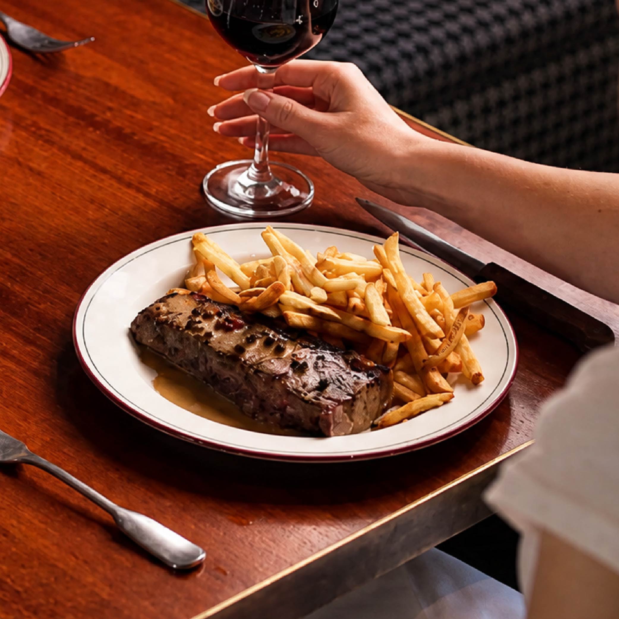 Sunday Roast - Plate with a serving of steak and French fries on a wooden table, with a person holding a glass of red wine in the background.