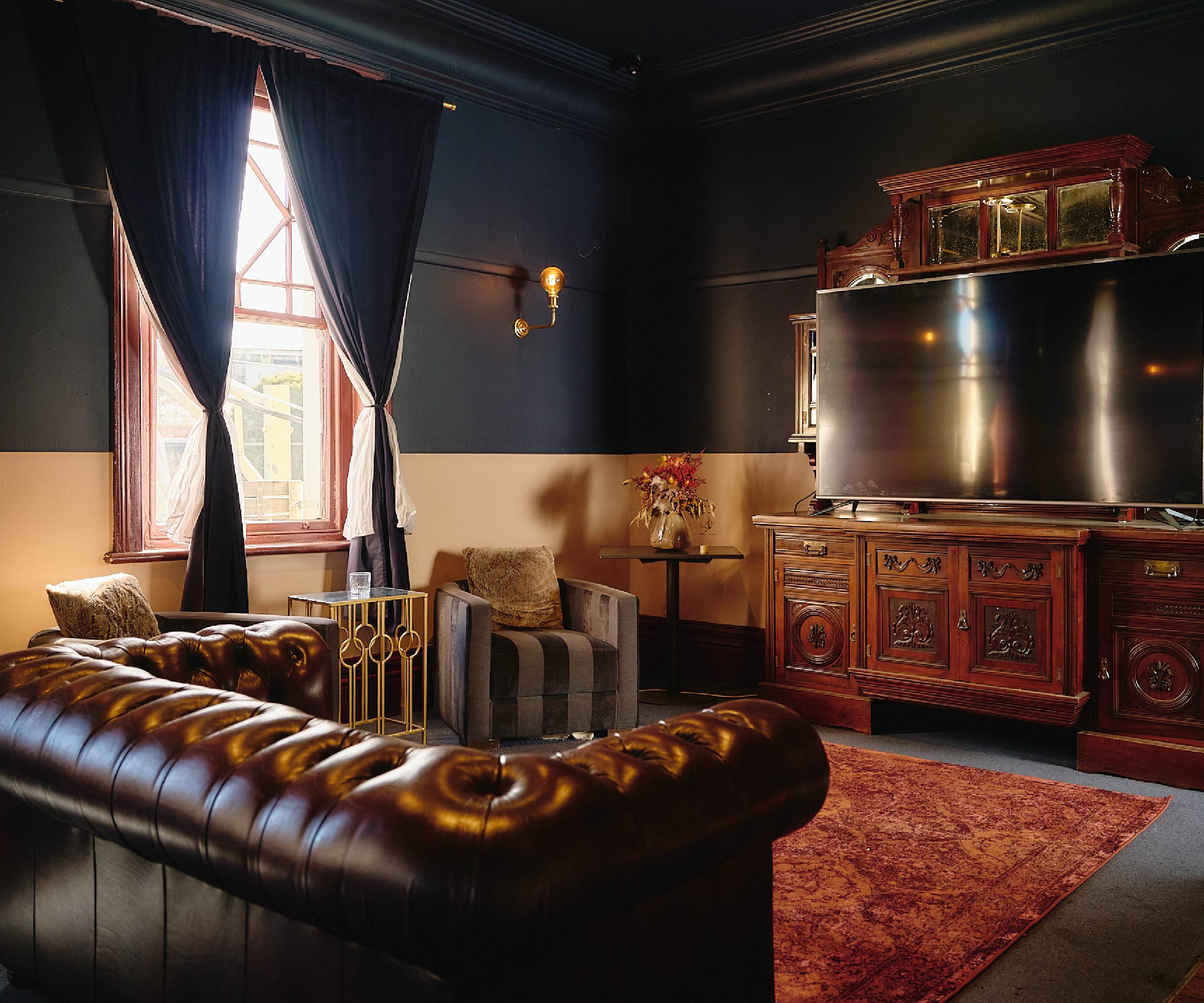 Cosy function room with dark walls, a large leather tufted sofa, a striped armchair with a pillow, a golden side table with a glass, a small table with a vase of dried flowers, a large flat-screen TV on an ornate wooden cabinet, and sunlight coming through a window with dark curtains.