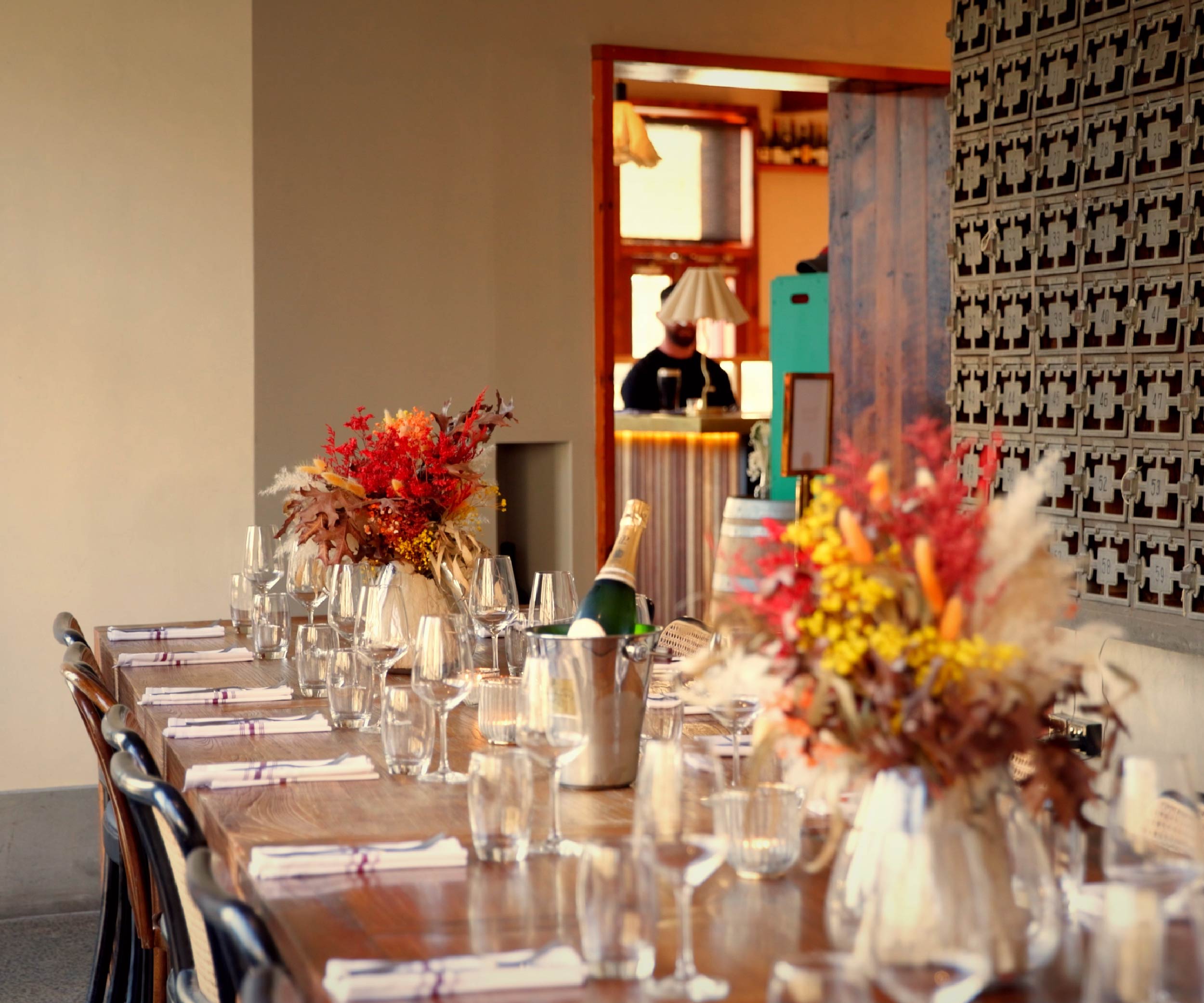 Elegant dining table set with white napkins, glassware, two floral centerpieces in warm autumn colors, and a champagne bottle in an ice bucket, with a bar area and a person blurred in the background.