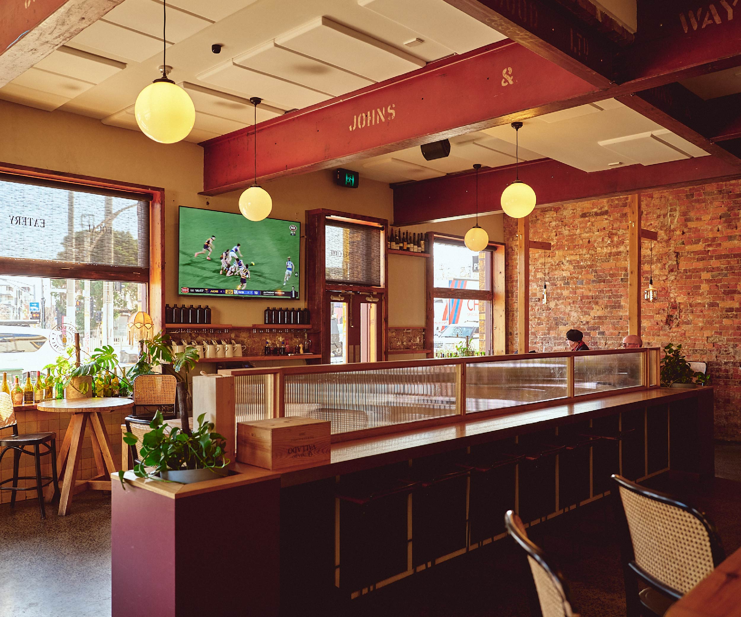 Interior of a cosy bar space with exposed brick walls, wooden beams, pendant globe lights, and a television showing a sports game, with plants and seating areas near windows.