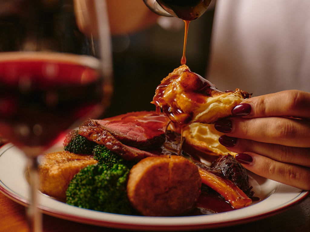 Person pouring brown gravy on a Yorkshire pudding held above a plate with sliced beef, roasted potatoes, broccoli, carrot, and a glass of red wine in the foreground.