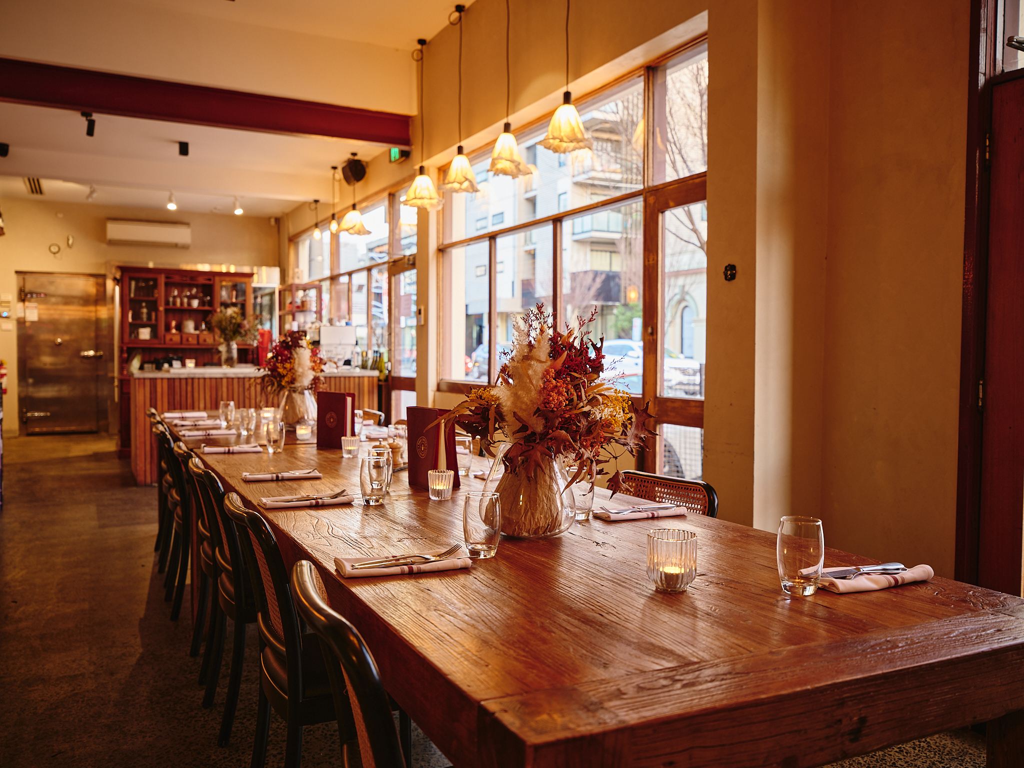 A woman with dark hair wearing a black jacket arranges a large vase of autumnal flowers on a long wooden dining table set with glasses, napkins, and silverware in a warmly lit, rustic restaurant interior.