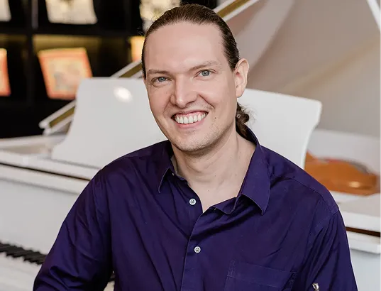Smiling man with long hair tied back wearing a purple shirt, seated in front of a white piano in a well-lit room.