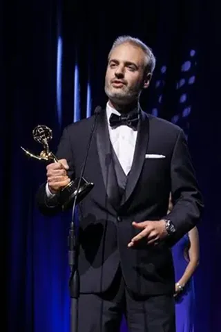 Man in a black tuxedo with a bow tie standing on stage holding an Emmy award statuette and speaking into a microphone against a dark blue curtain background.