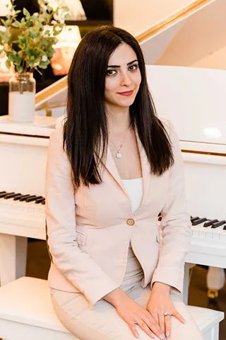Woman with long dark hair wearing a light beige blazer and skirt sitting on a white piano bench in front of a white grand piano, with a vase of flowers in the background.